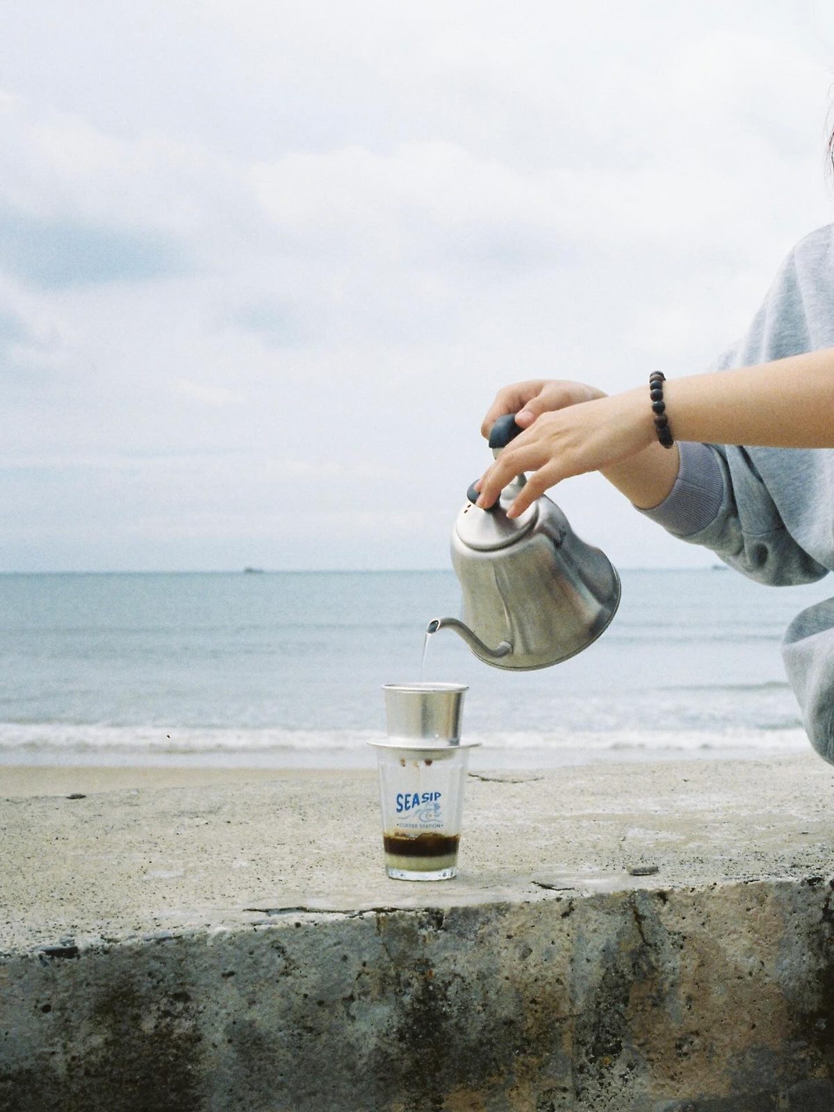 Ly cà phê đá mang thương hiệu Sea Sip trên bờ biển, cạnh ly pour-over trong bình thủy tinh