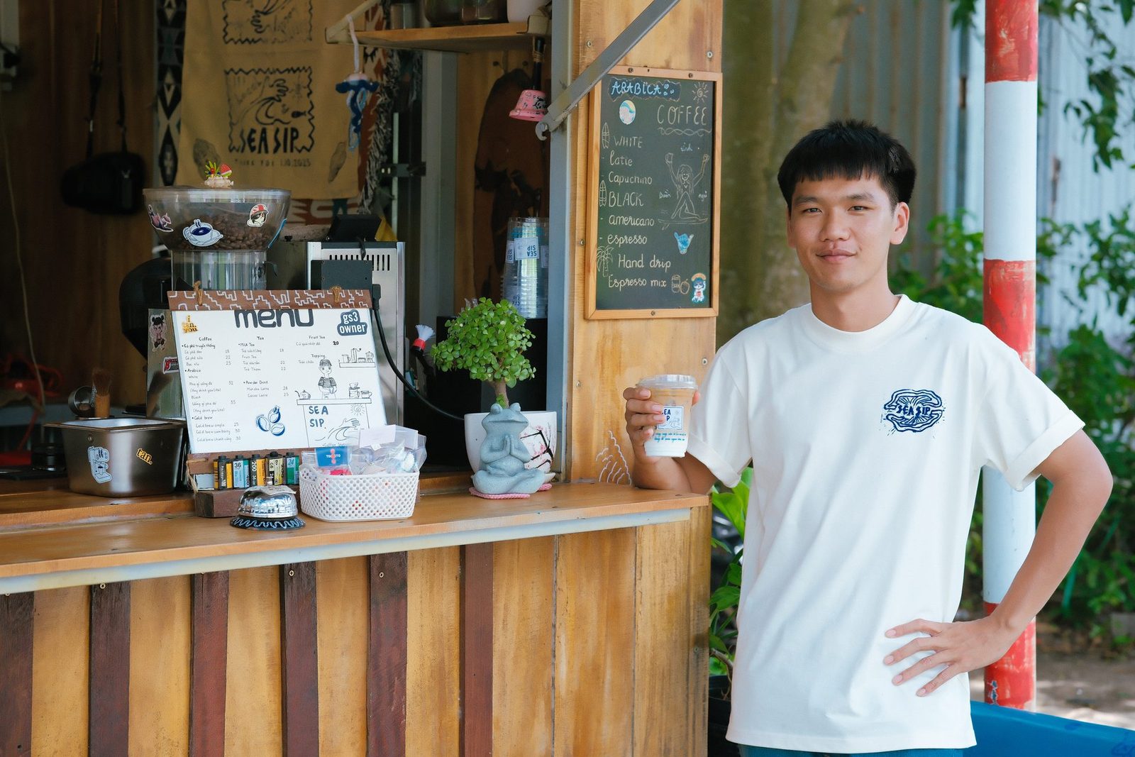 Quang đứng cạnh quầy với ly nước trên tay, bảng thực đơn phía sau