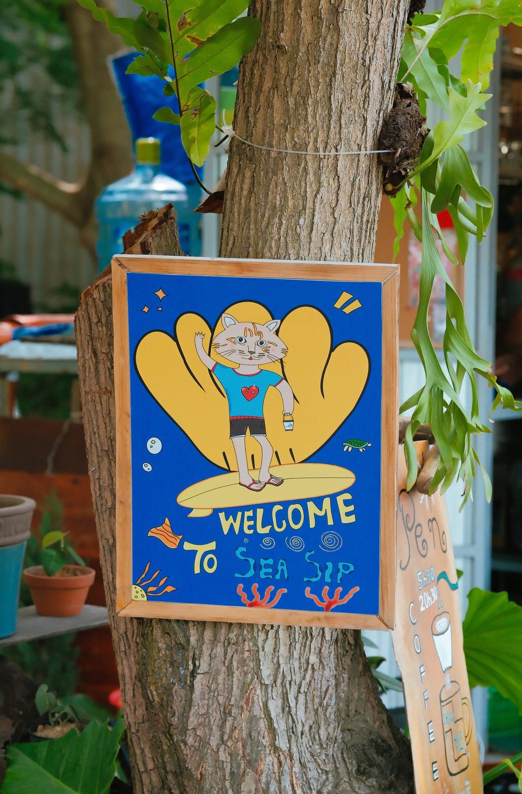 Bảng gỗ vẽ tay hình mèo lướt sóng, dòng chữ Welcome to Sea Sip đóng trên thân cây